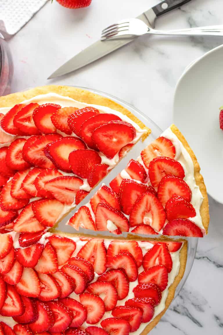 A strawberry tart with cream filling, reminiscent of a strawberry pizza, sits on a marble surface; one slice is separated, with a fork, knife, and plate nearby.