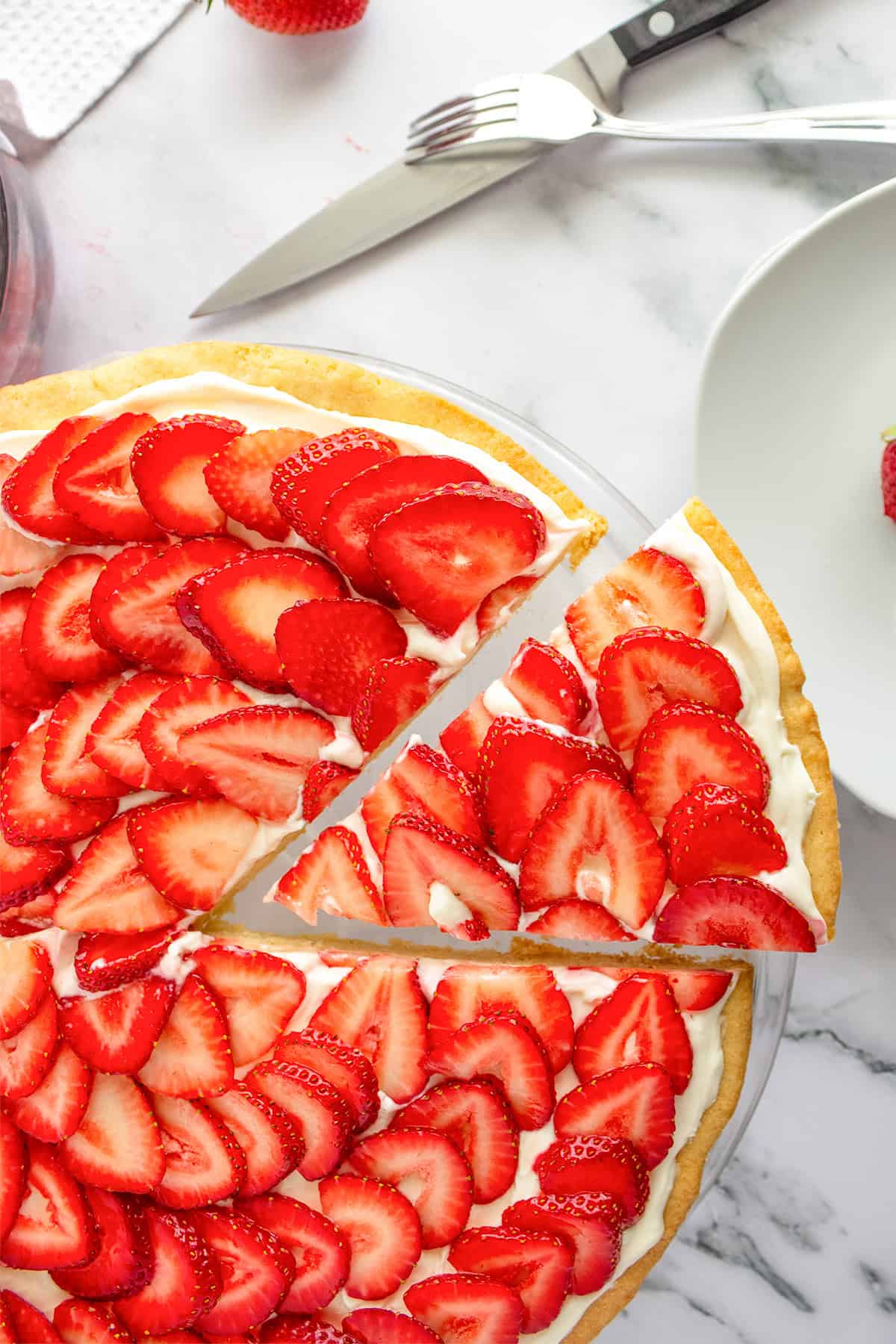 A strawberry tart with cream filling, reminiscent of a strawberry pizza, sits on a marble surface; one slice is separated, with a fork, knife, and plate nearby.