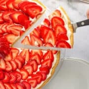A hand lifts a slice of strawberry tart—like a sweet strawberry pizza—topped with sliced strawberries and cream, with a fork and whole strawberries on a marble surface nearby.