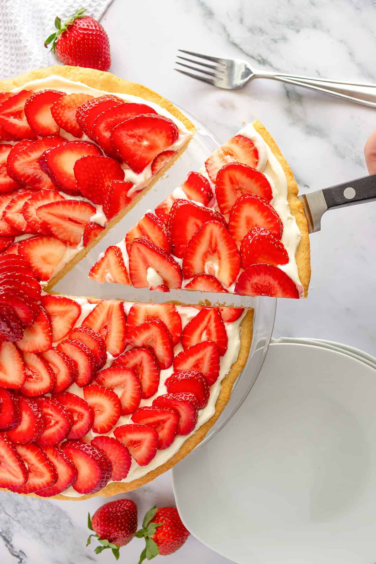 A hand lifts a slice of strawberry tart—like a sweet strawberry pizza—topped with sliced strawberries and cream, with a fork and whole strawberries on a marble surface nearby.