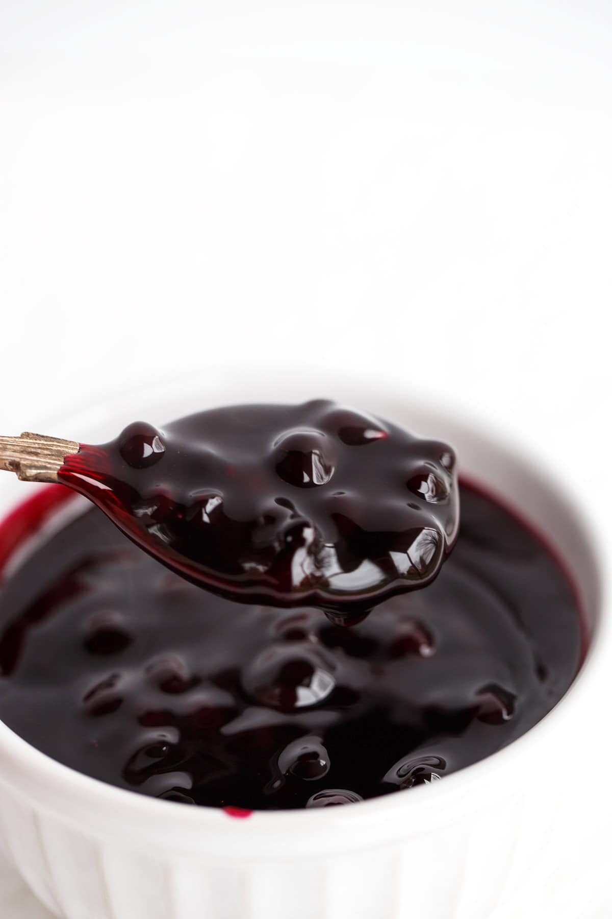 A spoon holding glossy, dark cherry pie filling above a white ramekin filled with the same filling, reminiscent in texture and shine to a homemade Blueberry Sauce, against a white background.