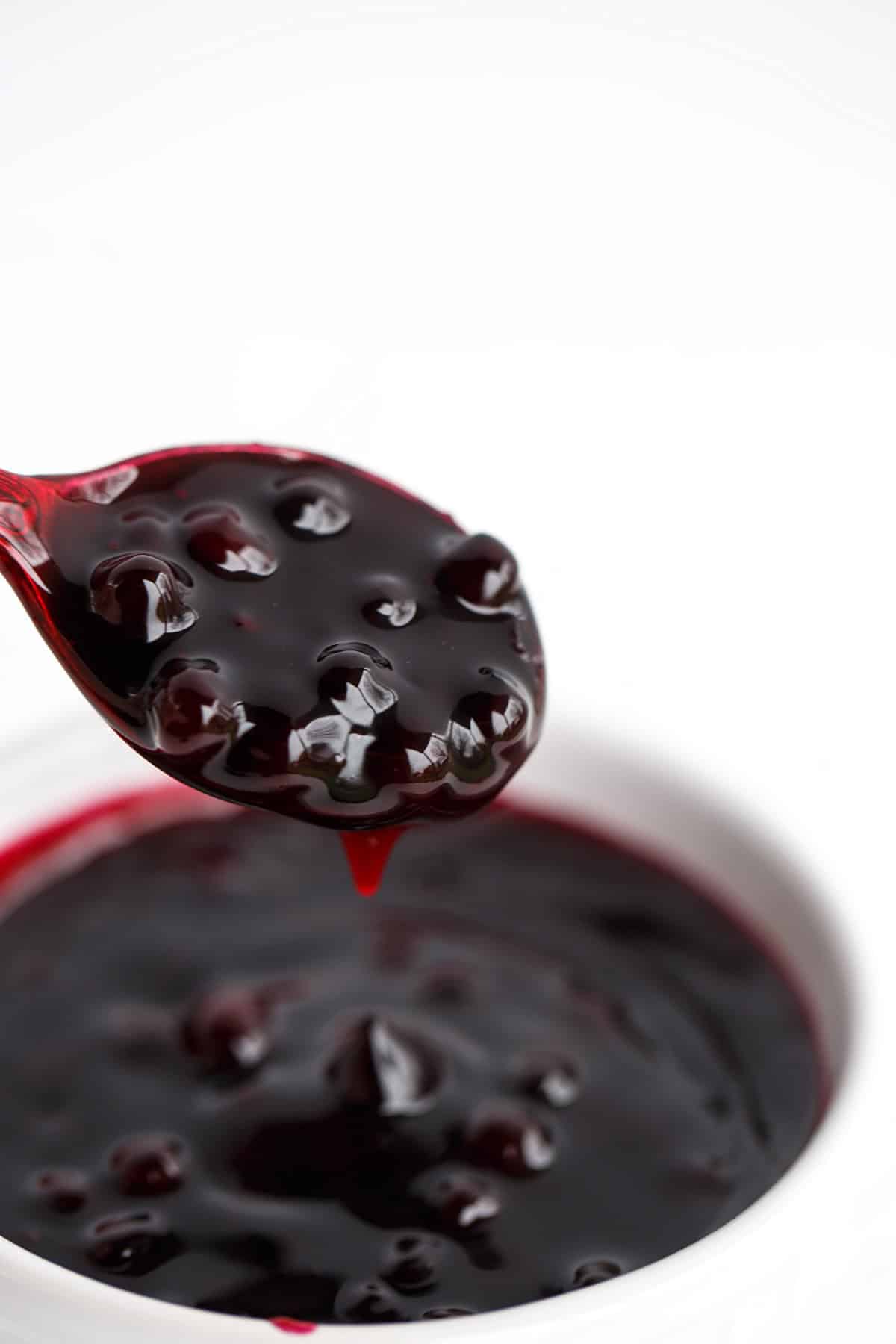 A spoon holding thick, dark blueberry sauce with whole berries hovers above a white bowl filled with more sauce, set against a clean white background.