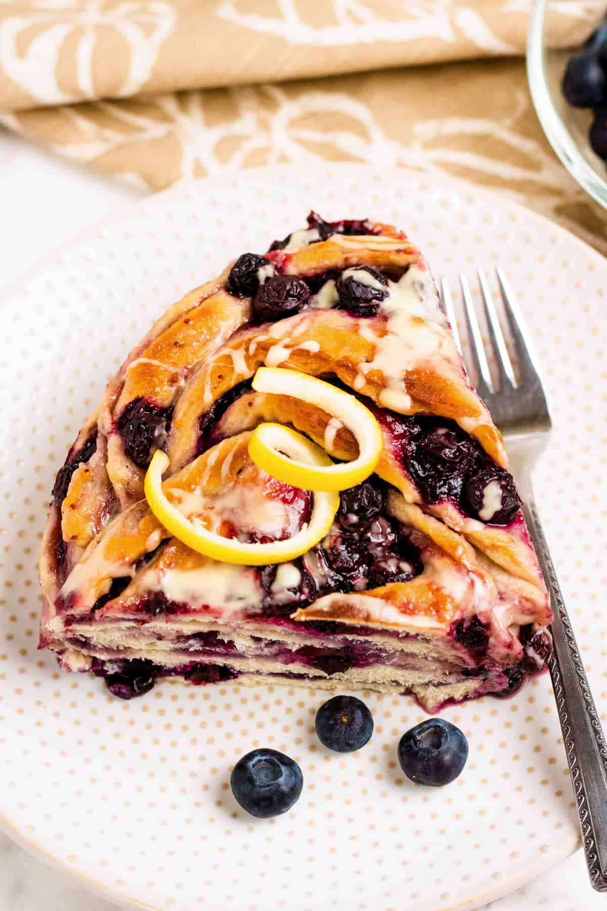 A slice of Blueberry Swirl Bread filled with blueberries, drizzled with icing, garnished with lemon zest, and served on a white plate with a fork and a few fresh blueberries.