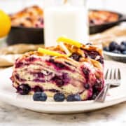 A slice of blueberry and lemon swirl bread sits on a white plate with fresh blueberries, a fork, and a bottle of milk in the background.