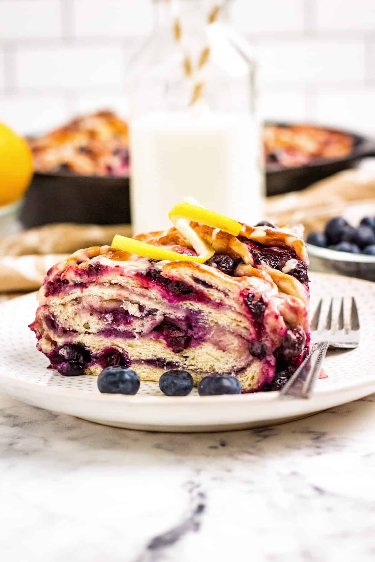 A slice of blueberry and lemon swirl bread sits on a white plate with fresh blueberries, a fork, and a bottle of milk in the background.
