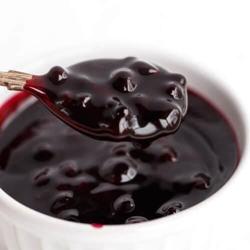 A spoonful of glossy, dark blueberry sauce is held above a white ramekin filled with the same sauce.