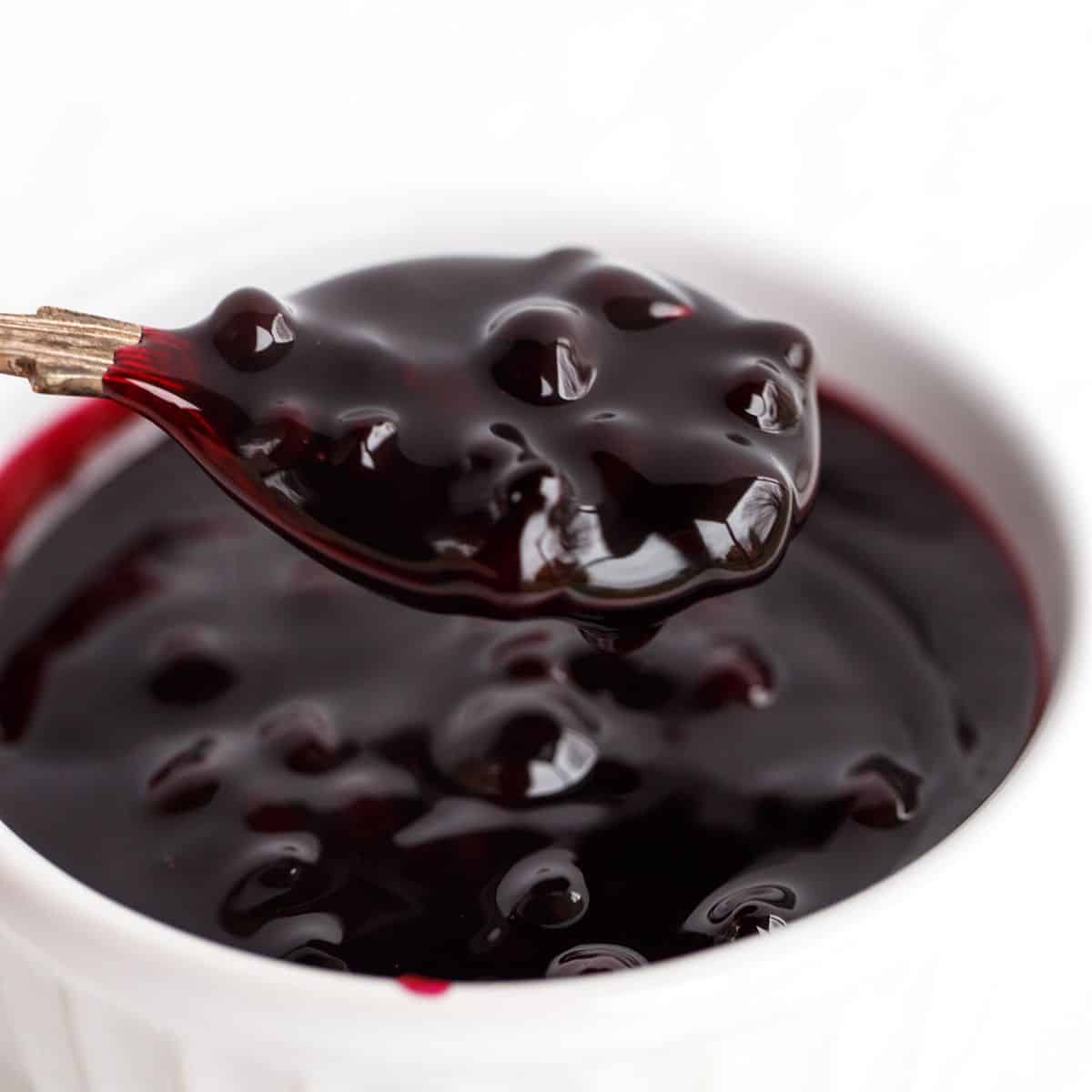 A spoonful of glossy, dark blueberry sauce is held above a white ramekin filled with the same sauce.