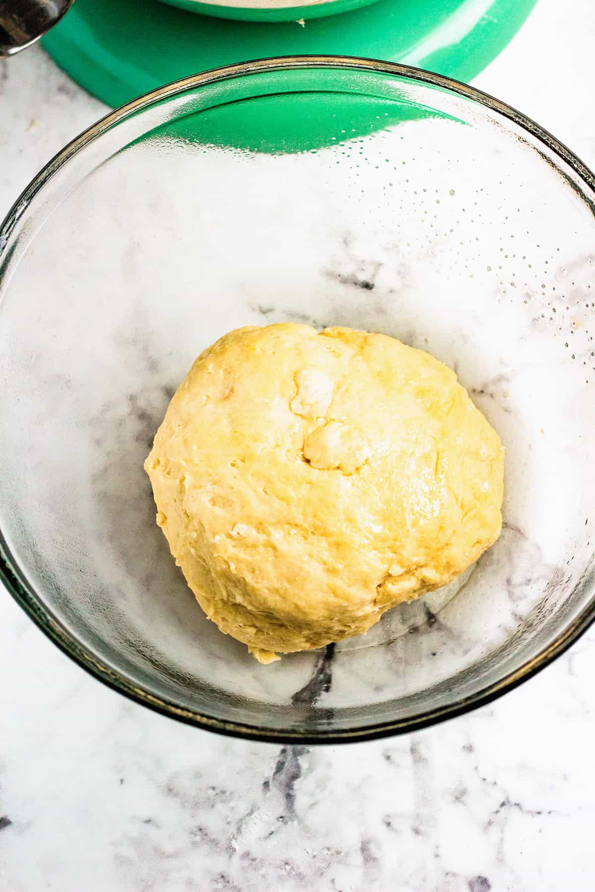 A ball of dough for Blueberry Swirl Bread rests in a clear glass bowl on a white marble surface, with part of a green kitchen appliance visible in the background.