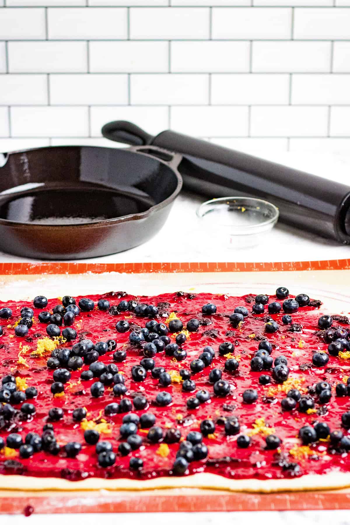 Rolled-out dough ready for Blueberry Swirl Bread, topped with berry filling and fresh blueberries on a silicone mat, with a skillet, rolling pin, and small glass bowl in the background.