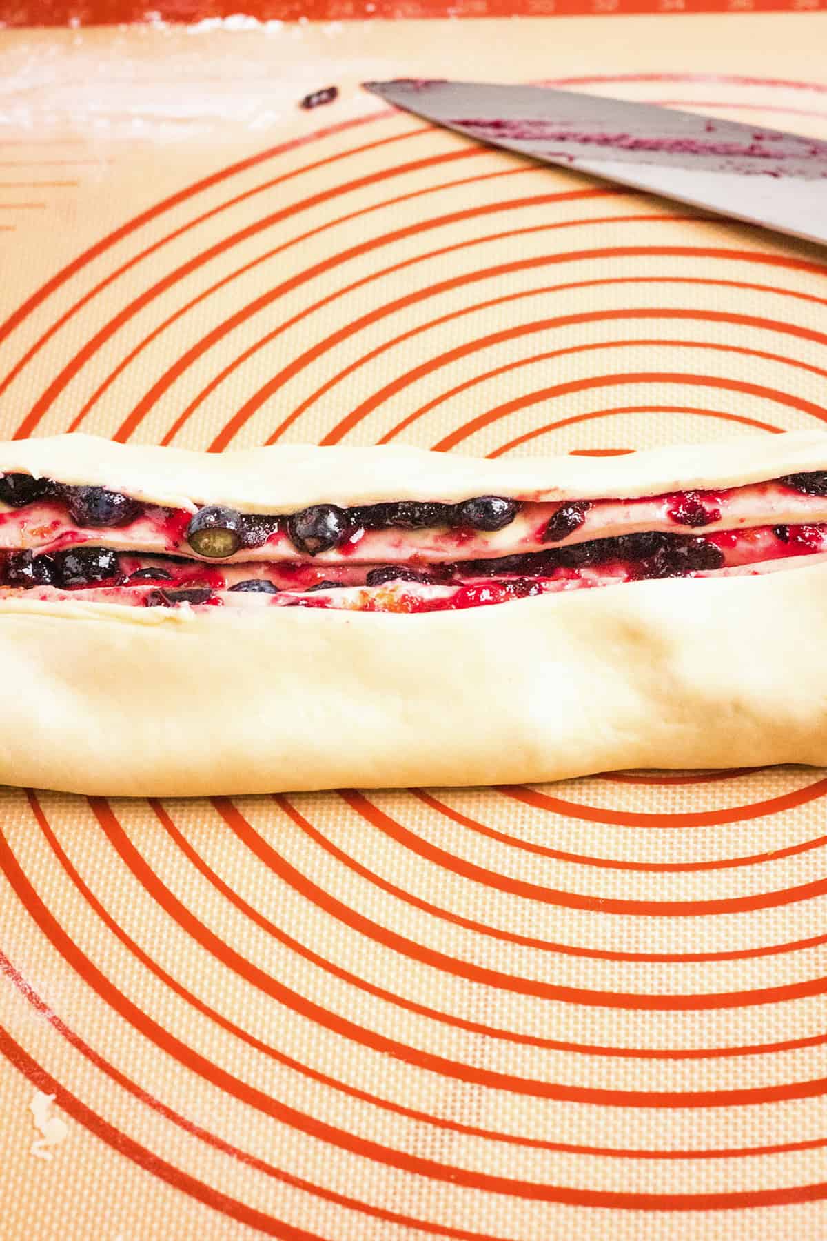 Unbaked pastry dough filled with a blueberry swirl bread mixture is partially rolled on a silicone baking mat with red concentric circles and a pastry cutter nearby.