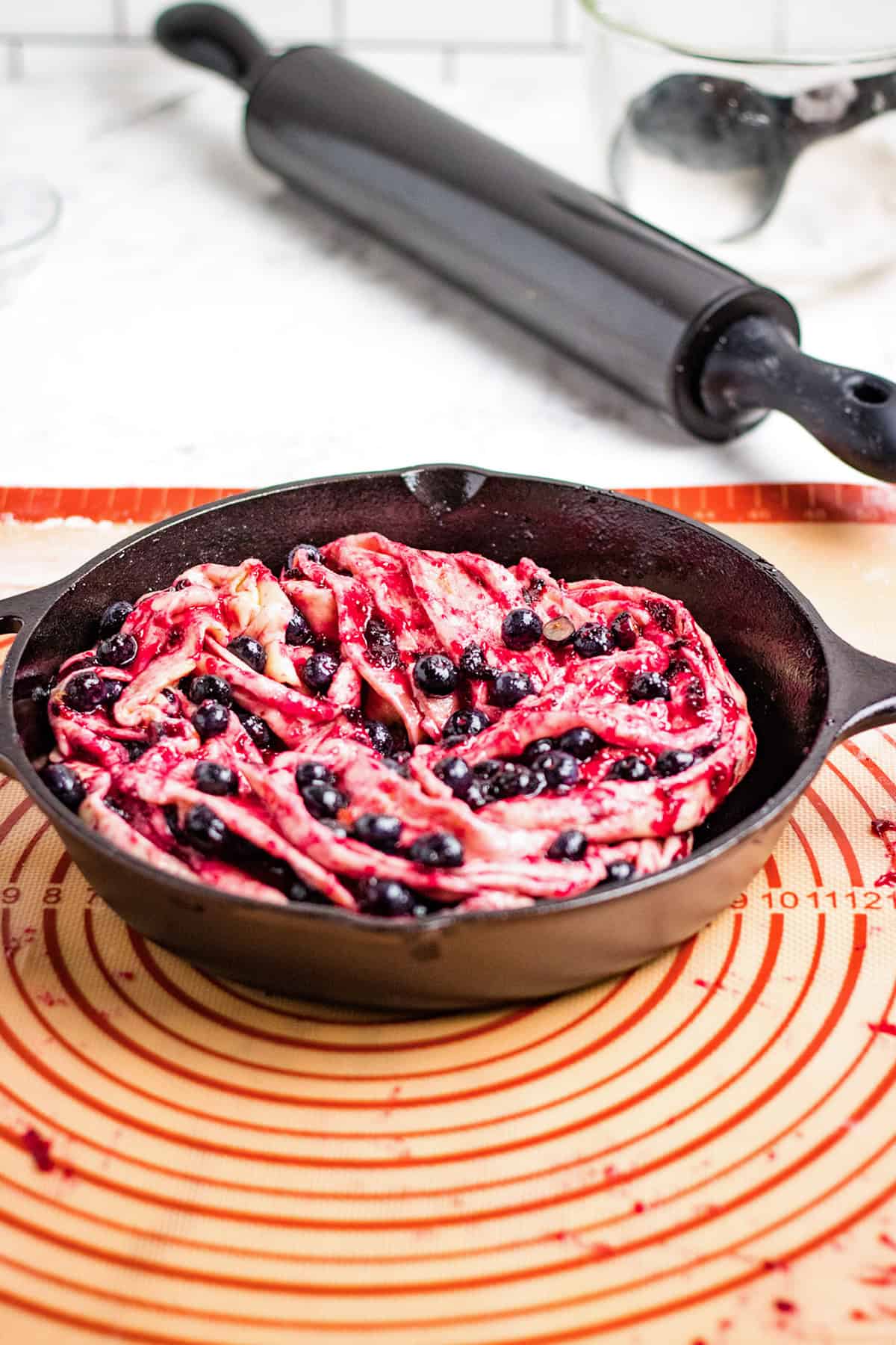 Unbaked Blueberry Swirl Bread dough, twisted and filled with berries, rests in a cast iron skillet on a silicone baking mat; a rolling pin and mixing bowl linger in the background.
