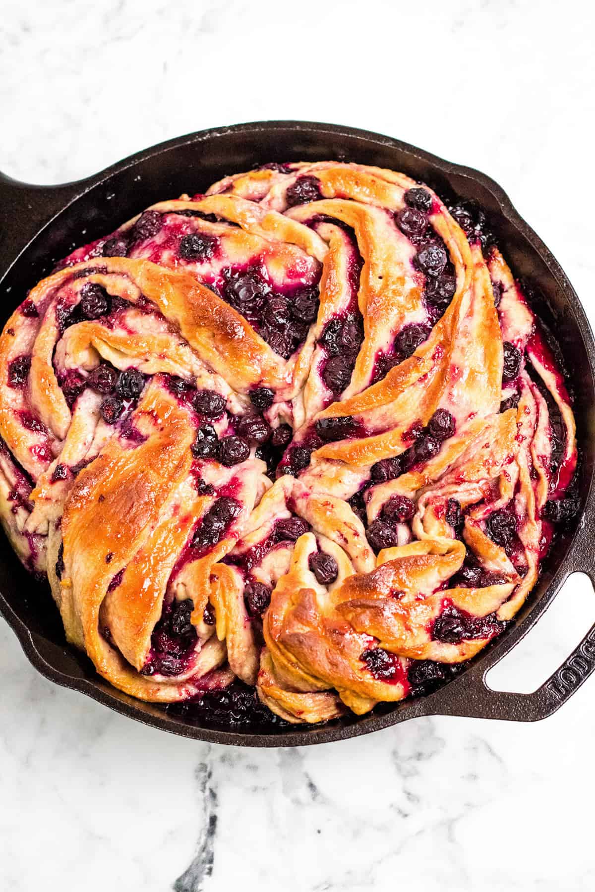 Blueberry Swirl Bread with a golden-brown crust, twisted to perfection and baked in a cast iron skillet on a white marble surface.