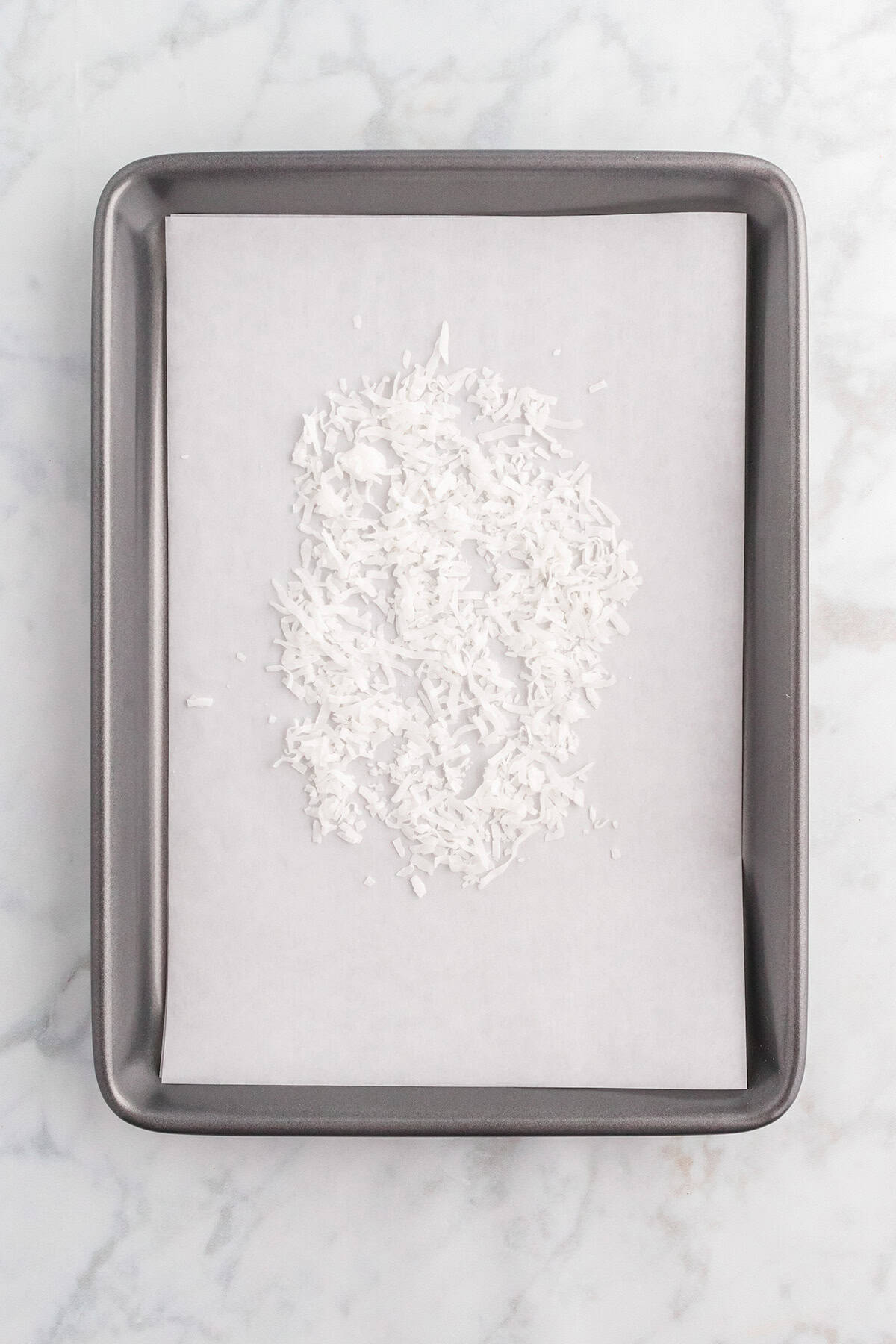 A baking sheet lined with parchment paper topped with a small pile of shredded coconut, set on a marble countertop—perfectly prepped for adding the signature coconut crunch to your Million Dollar Pie.