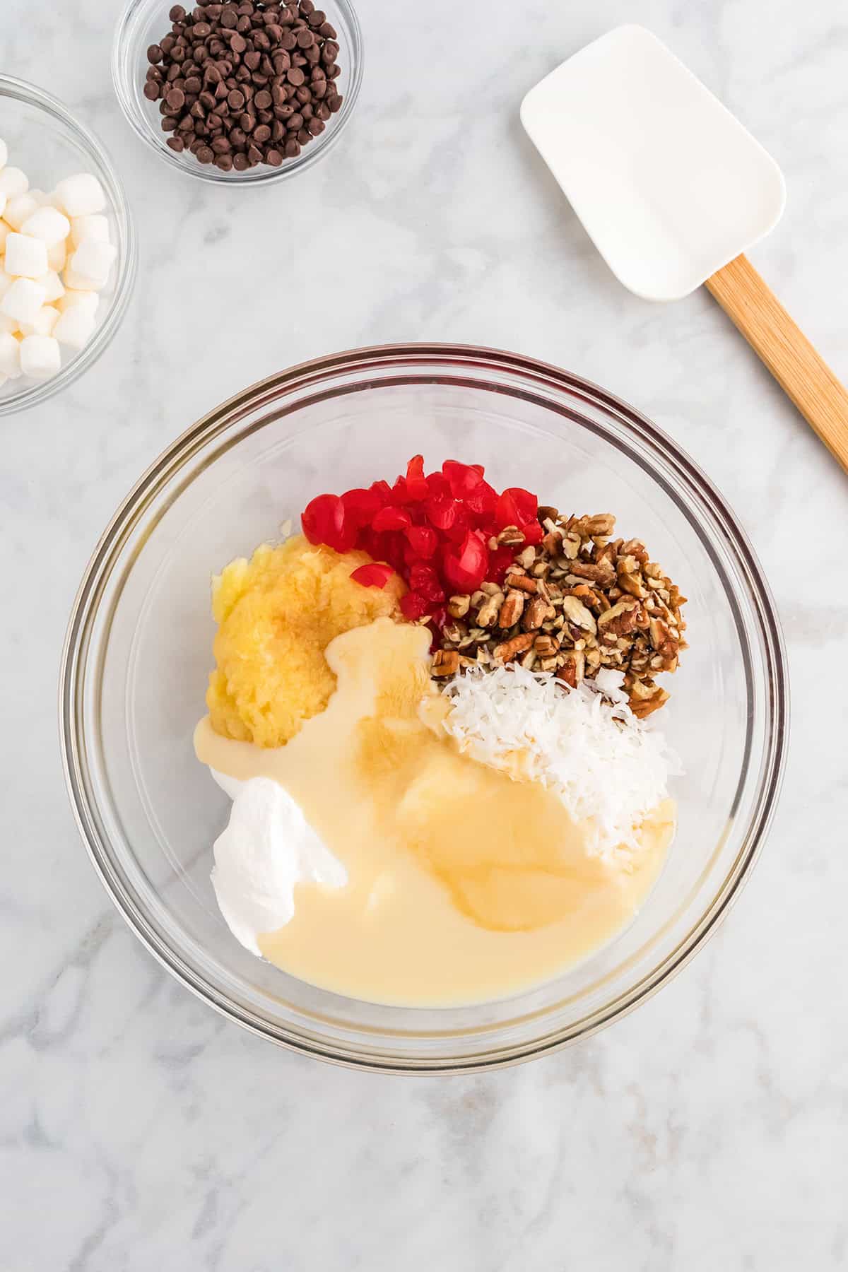 A glass bowl with pineapple, cherries, pecans, coconut, and sweetened condensed milk—classic ingredients for a delicious Million Dollar Pie. Nearby are bowls of mini marshmallows and chocolate chips, plus a spatula on a marble countertop.