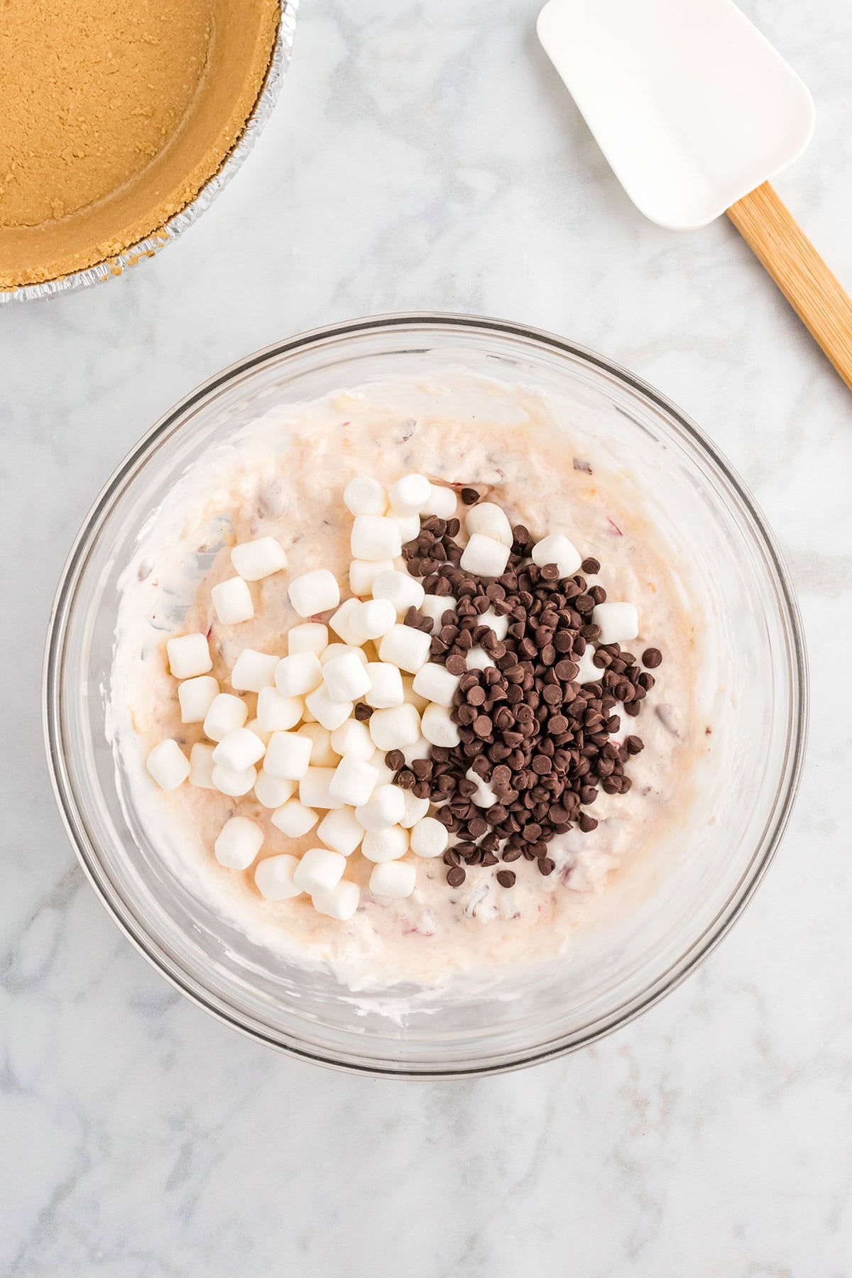A glass bowl contains a creamy Million Dollar Pie mixture with mini marshmallows and chocolate chips on top, next to a spatula and a pie crust.