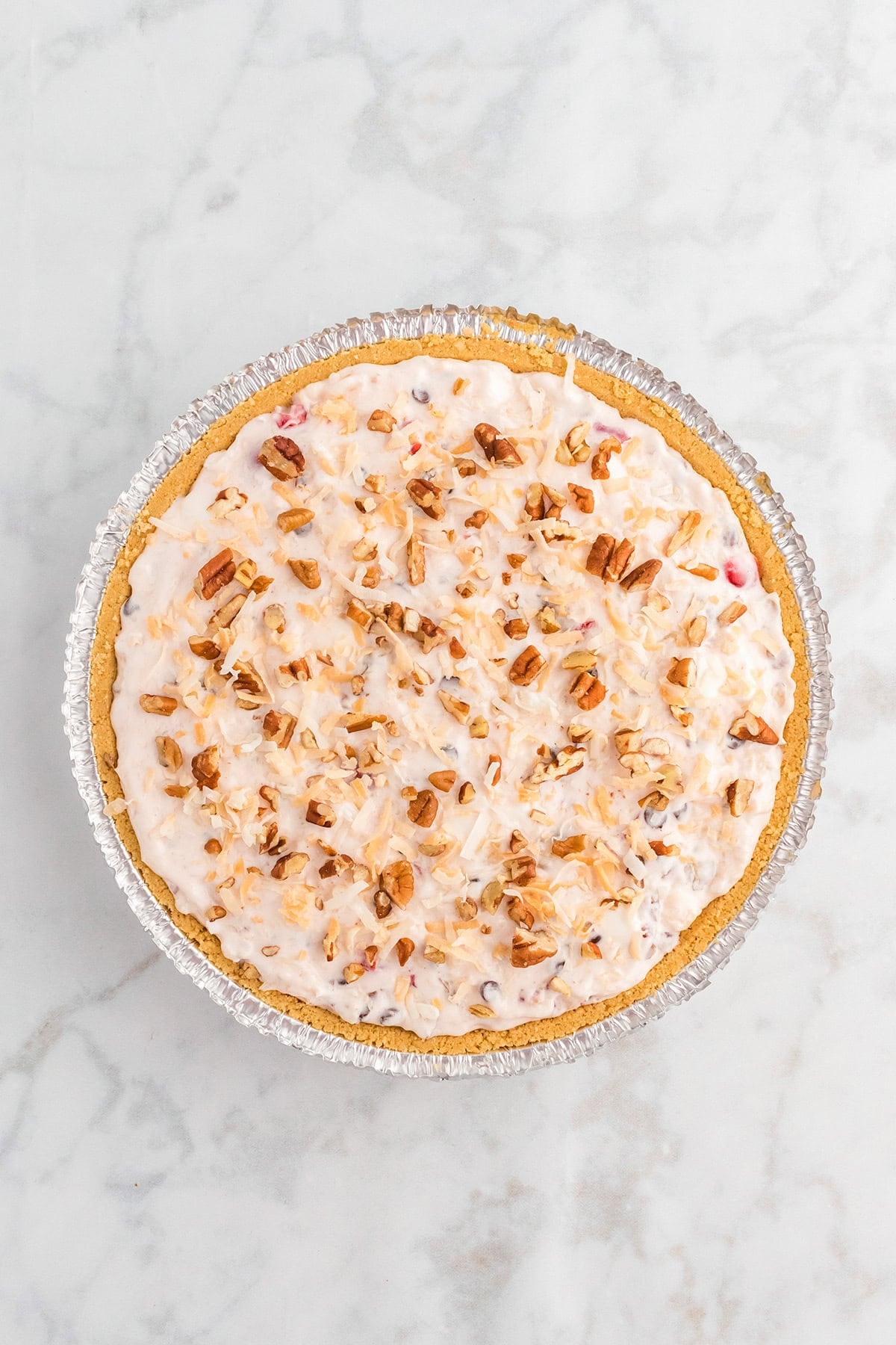 A whole Million Dollar Pie with a graham cracker crust, topped with a creamy filling and sprinkled with chopped nuts, shown from above on a marble surface.