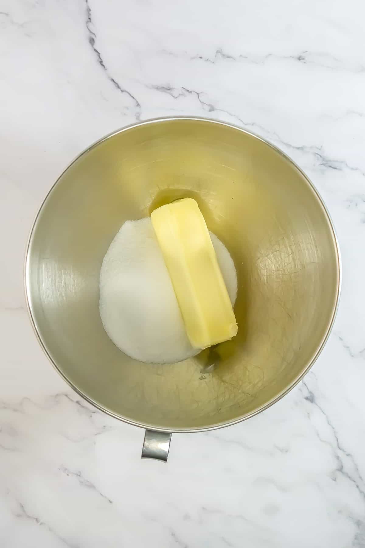 A metal mixing bowl contains a stick of butter and a mound of granulated sugar, set on a white marble surface—ingredients ready to be transformed into a delicious Raspberry Cake.