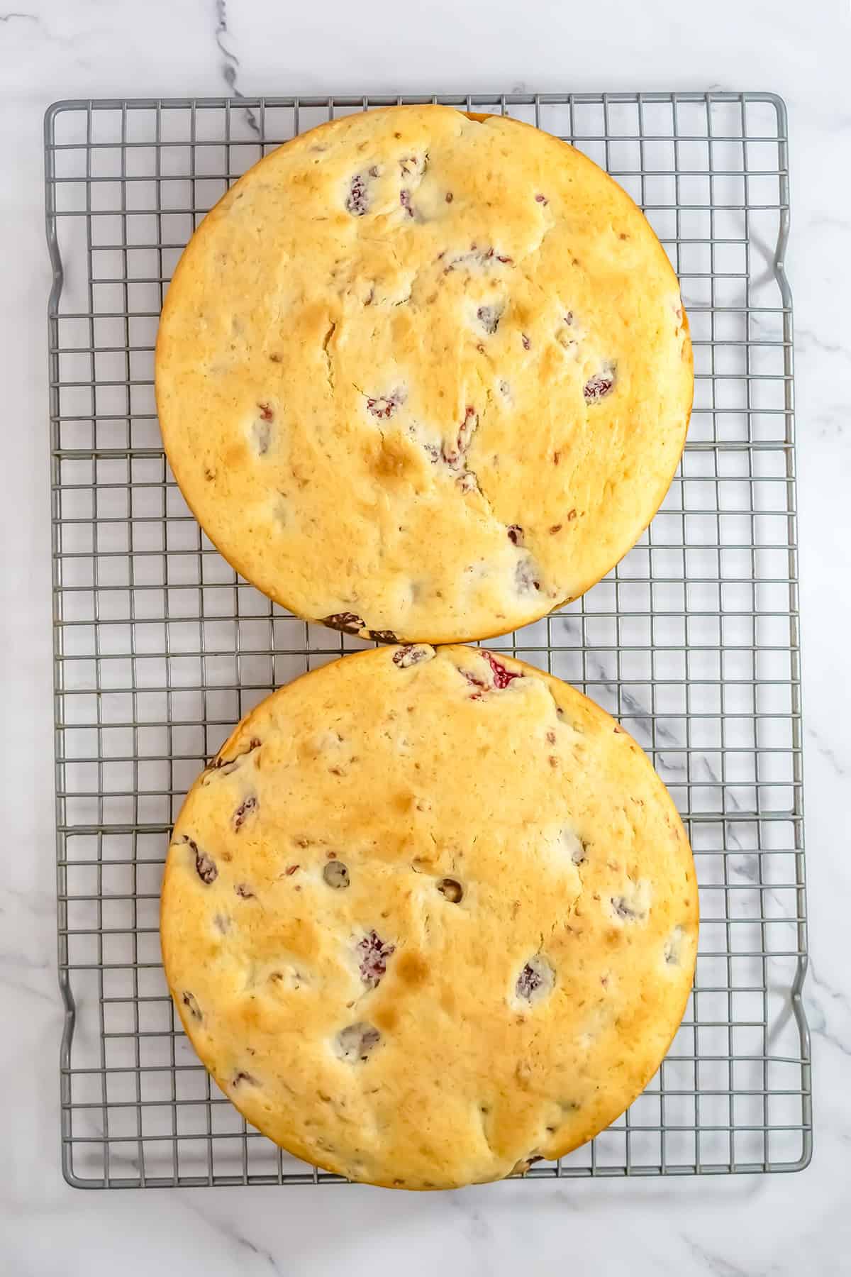 Two round, golden-brown raspberry cakes with visible fruit pieces cool on a wire rack placed on a white marble surface.