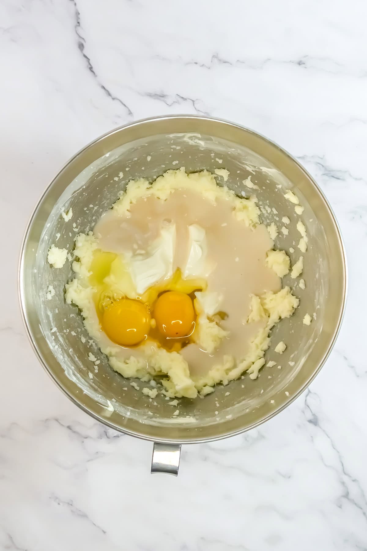 A metal mixing bowl contains creamed butter and sugar, two raw eggs, and liquid sweetened condensed milk on a white marble surface—ingredients ready to be transformed into a delicious Raspberry Cake.