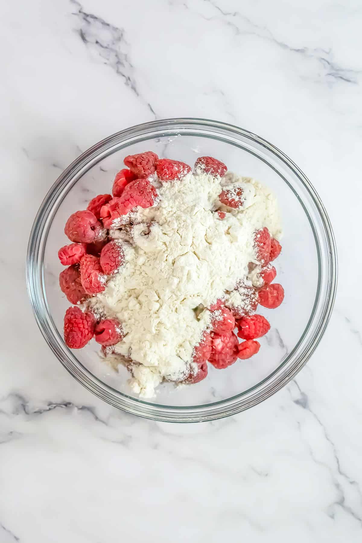 A glass bowl containing raspberries and flour sits on a white marble surface, ready to be transformed into a delicious Raspberry Cake.