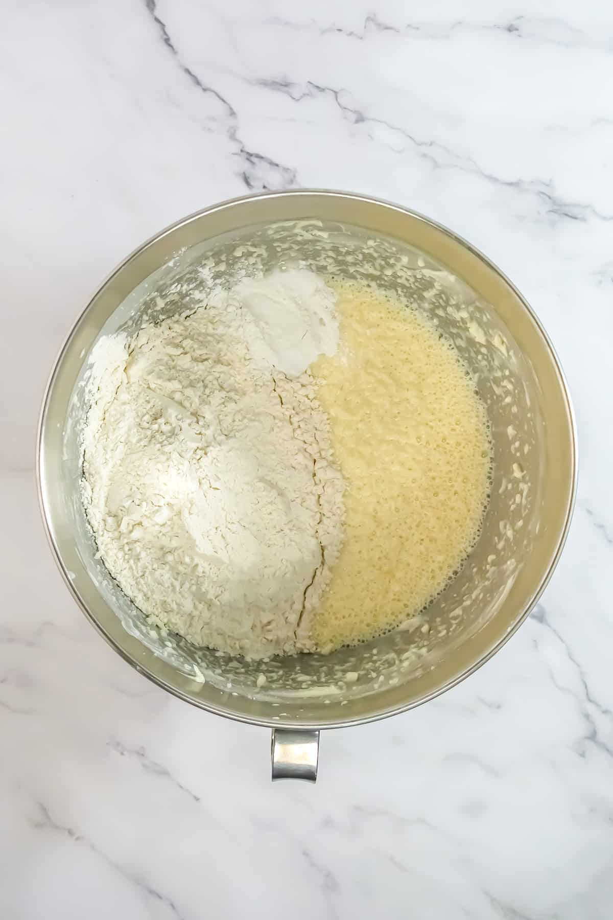 A metal mixing bowl containing flour and a wet batter mixture for a delicious Raspberry Cake, viewed from above on a white marble surface.