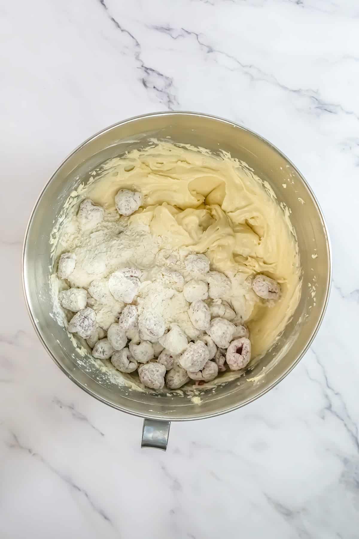 Metal mixing bowl with thick yellow batter, coated fresh blueberries, and flour on a white marble surface—ingredients ready to be mixed for baking a delicious Raspberry Cake.