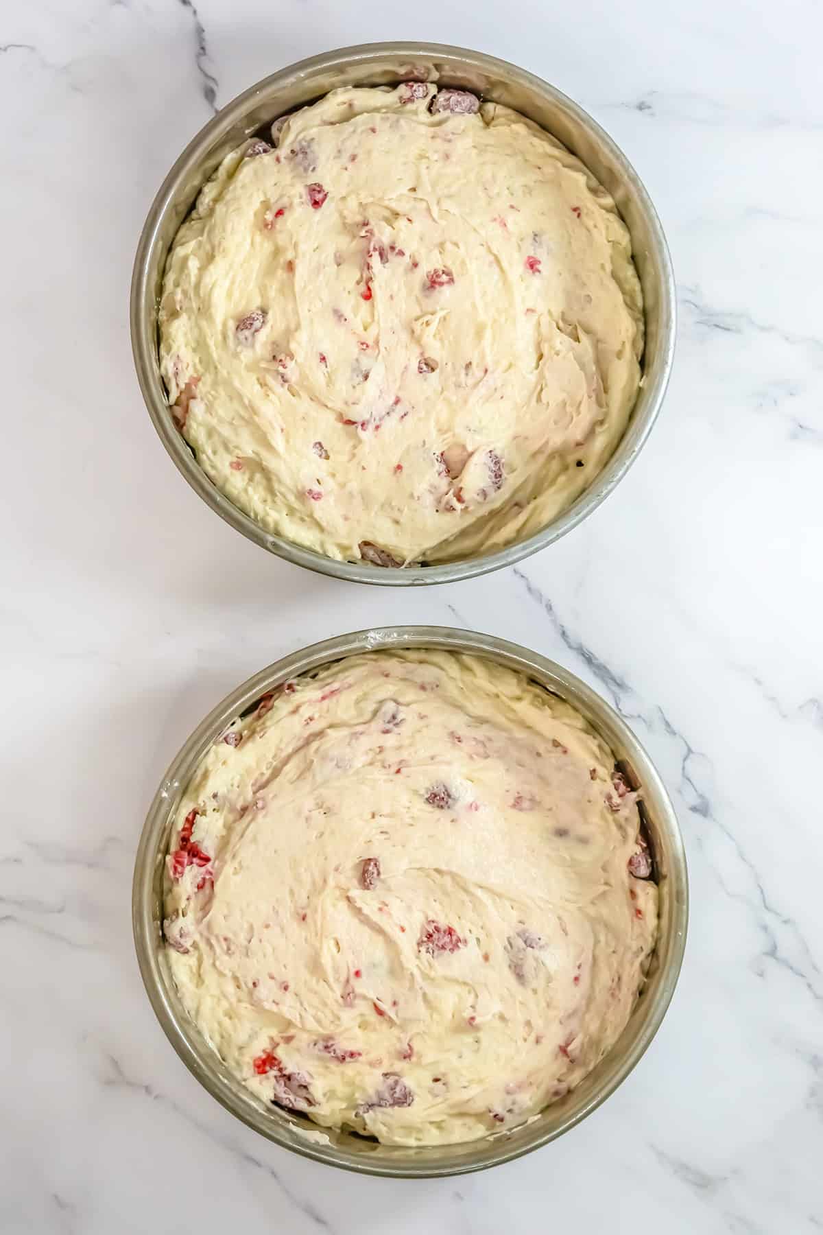 Two round cake pans filled with unbaked Raspberry Cake batter, mixed with visible red fruit pieces, are placed on a white marble surface.