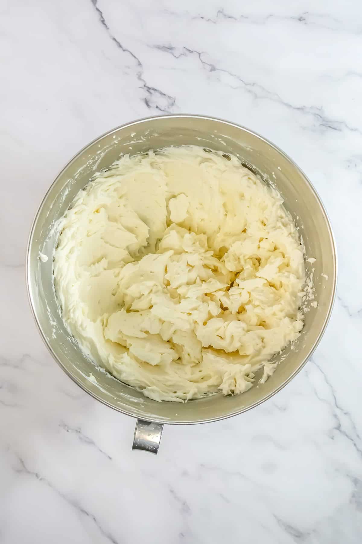 A metal mixing bowl filled with whipped cream cheese frosting sits on a white marble surface, ready to top a luscious Raspberry Cake.