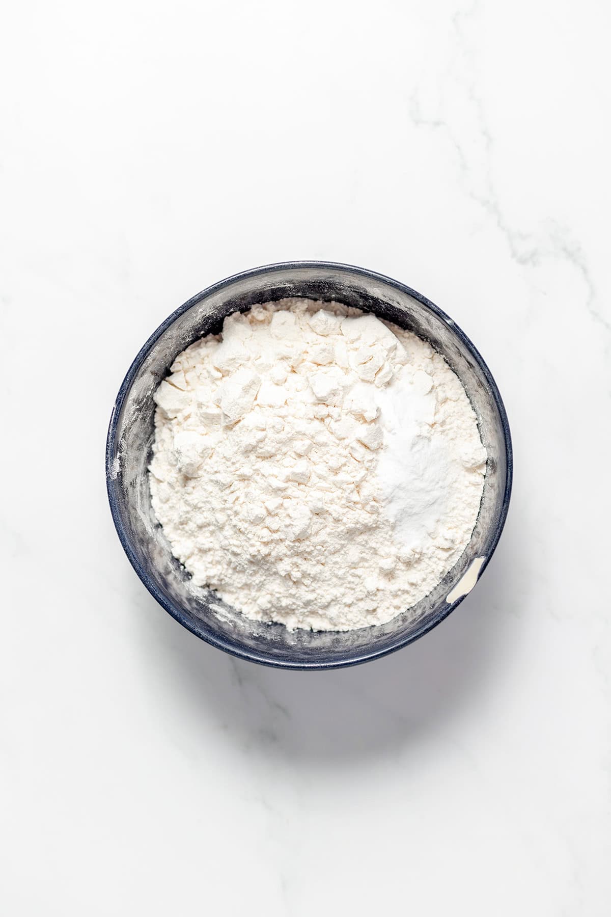 A black bowl filled with white flour, perfect for baking white chocolate raspberry cookies, sits on a white marble surface, viewed from above.