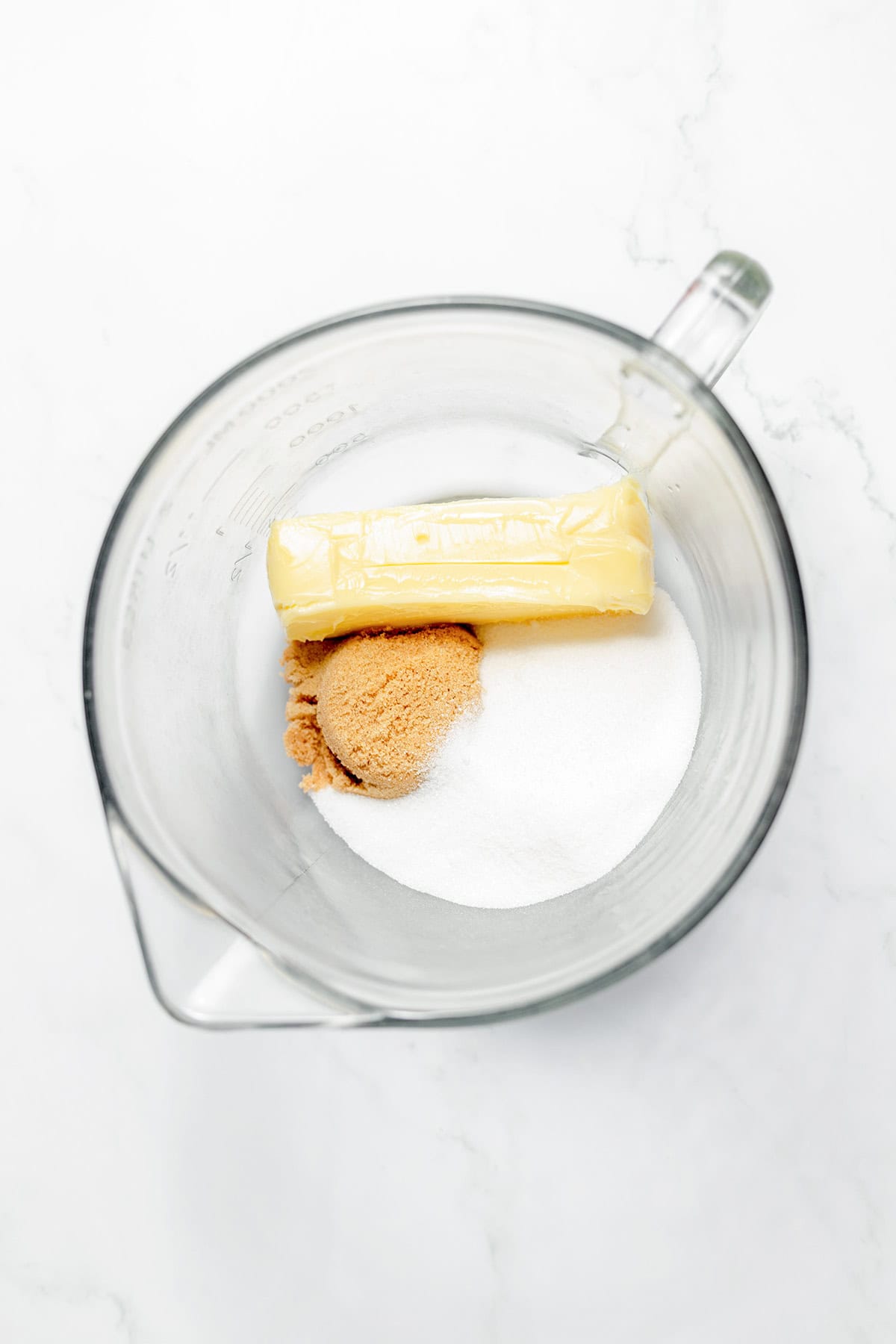 A glass mixing bowl containing a stick of butter, a mound of brown sugar, and a mound of granulated white sugar on a white surface—essential ingredients for delicious White Chocolate Cookies.