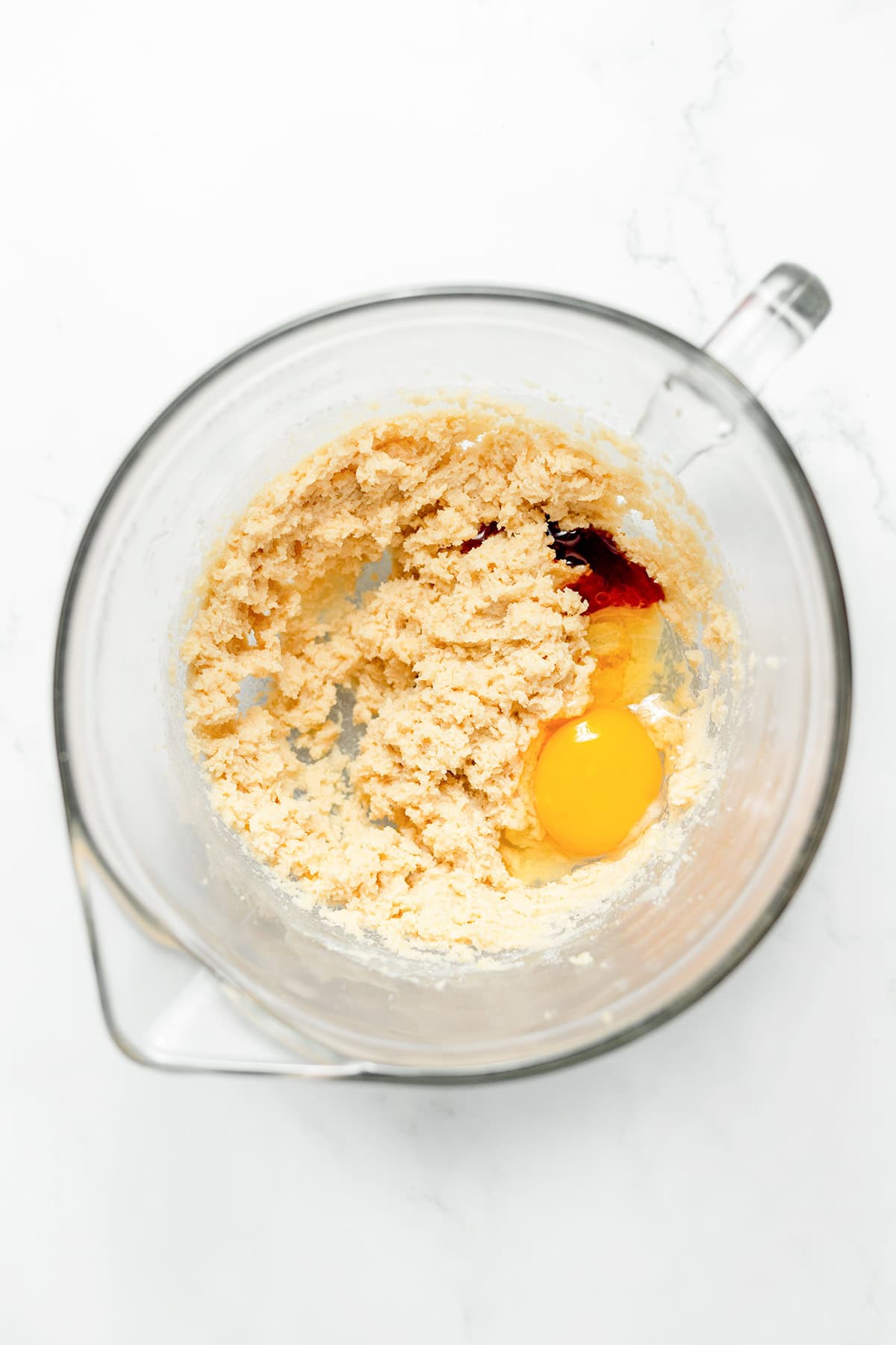 A glass mixing bowl with creamed butter and sugar, an egg, and vanilla extract on a white surface—perfect for starting a batch of homemade White Chocolate Cookies or irresistible Raspberry Cookies.