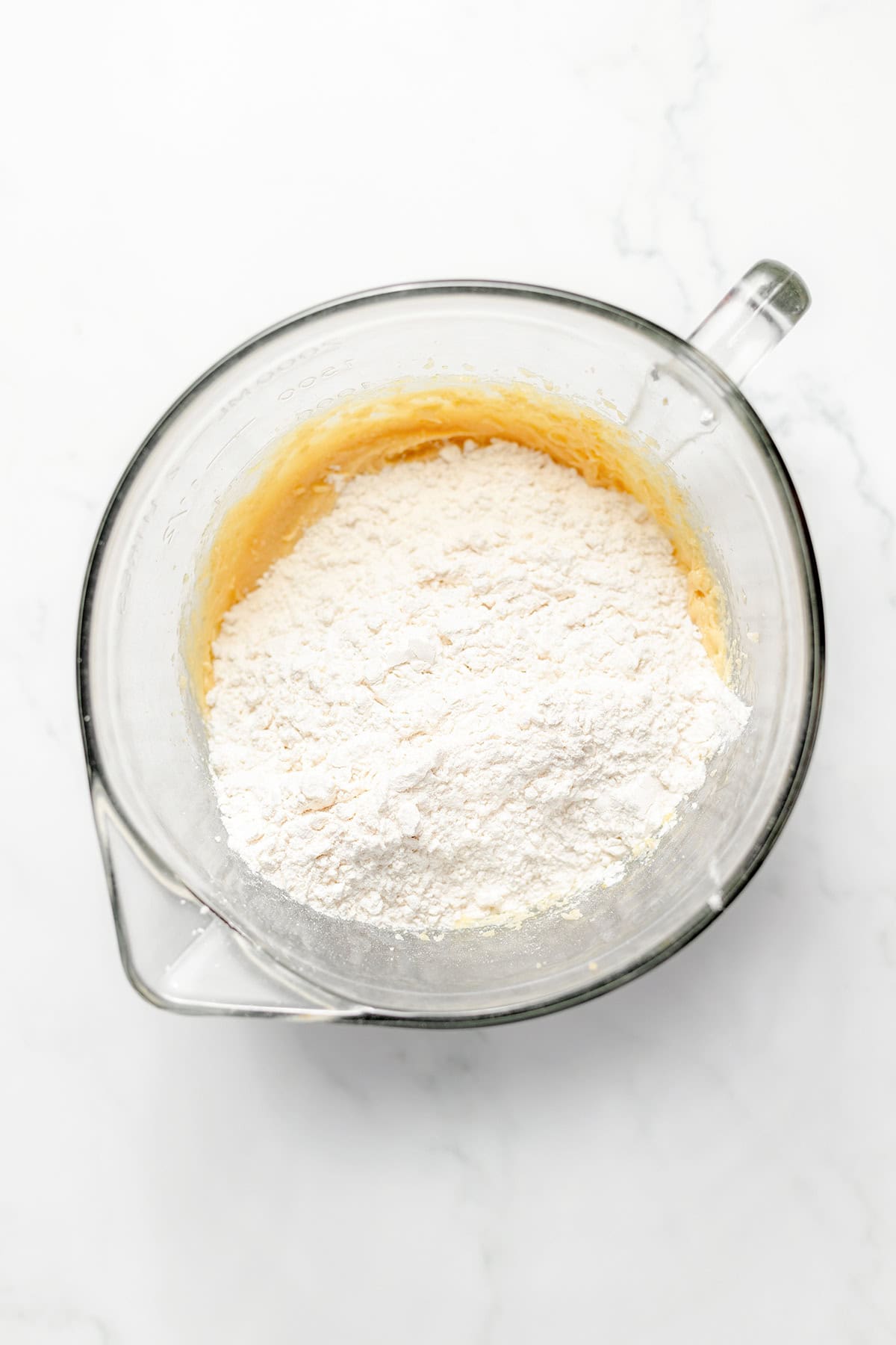 Clear glass mixing bowl containing flour on top of a yellow batter for white chocolate cookies, sitting on a white marble surface.