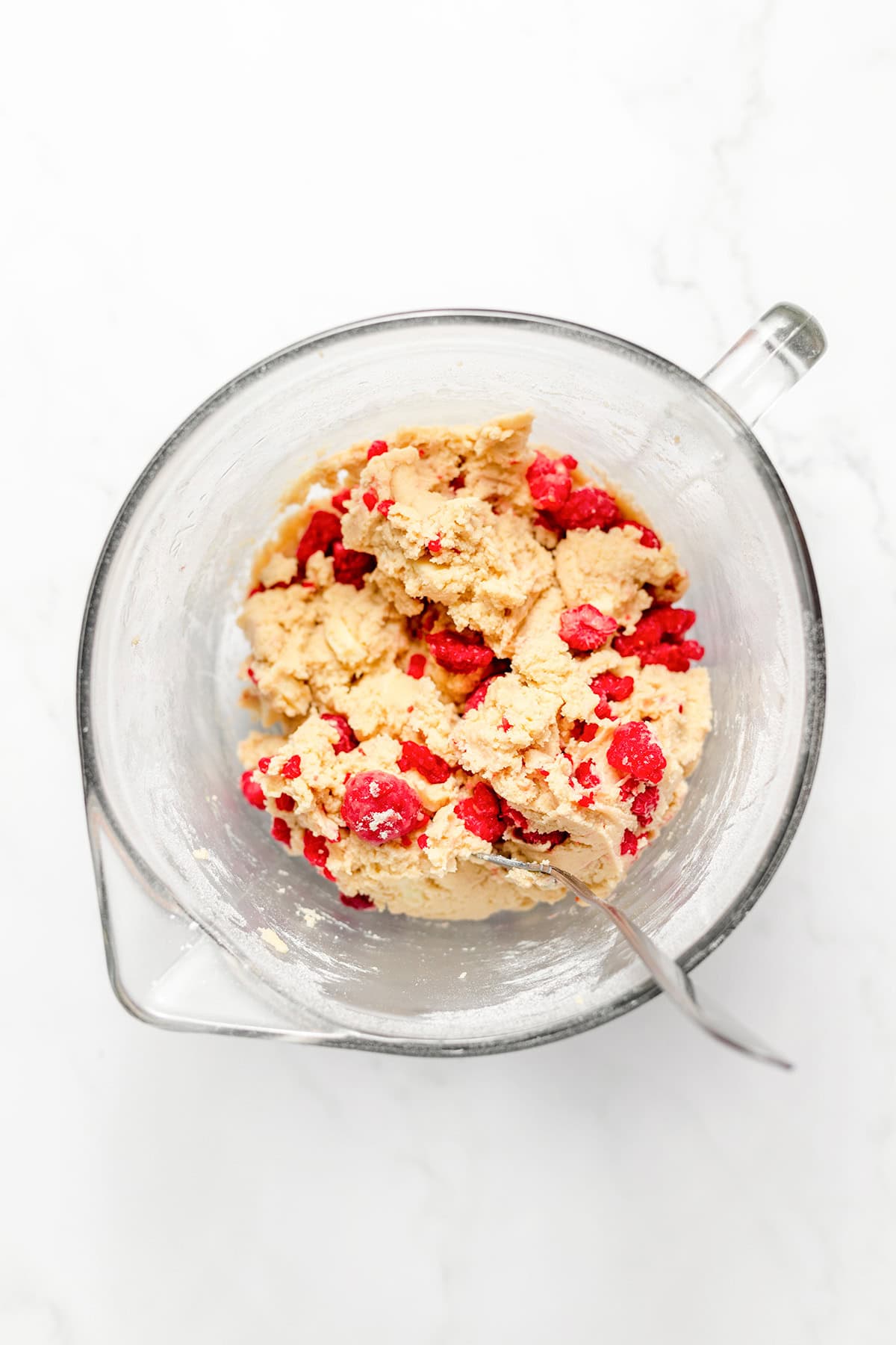 A glass mixing bowl containing raspberry cookie dough with visible red pieces and chunks of white chocolate, and a metal spoon resting inside, on a white marble surface—perfect for making delicious White Chocolate Raspberry Cookies.