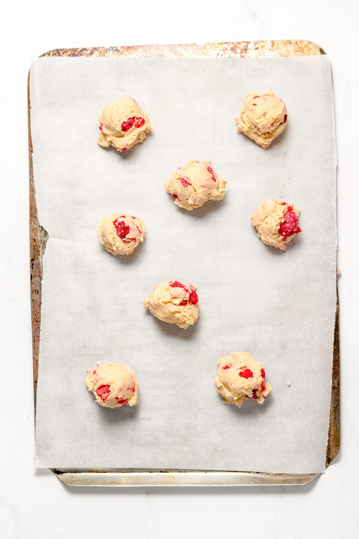 Eight scoops of cookie dough with red raspberry bits are arranged on a parchment-lined baking sheet, ready to be baked into delicious White Chocolate Raspberry Cookies.