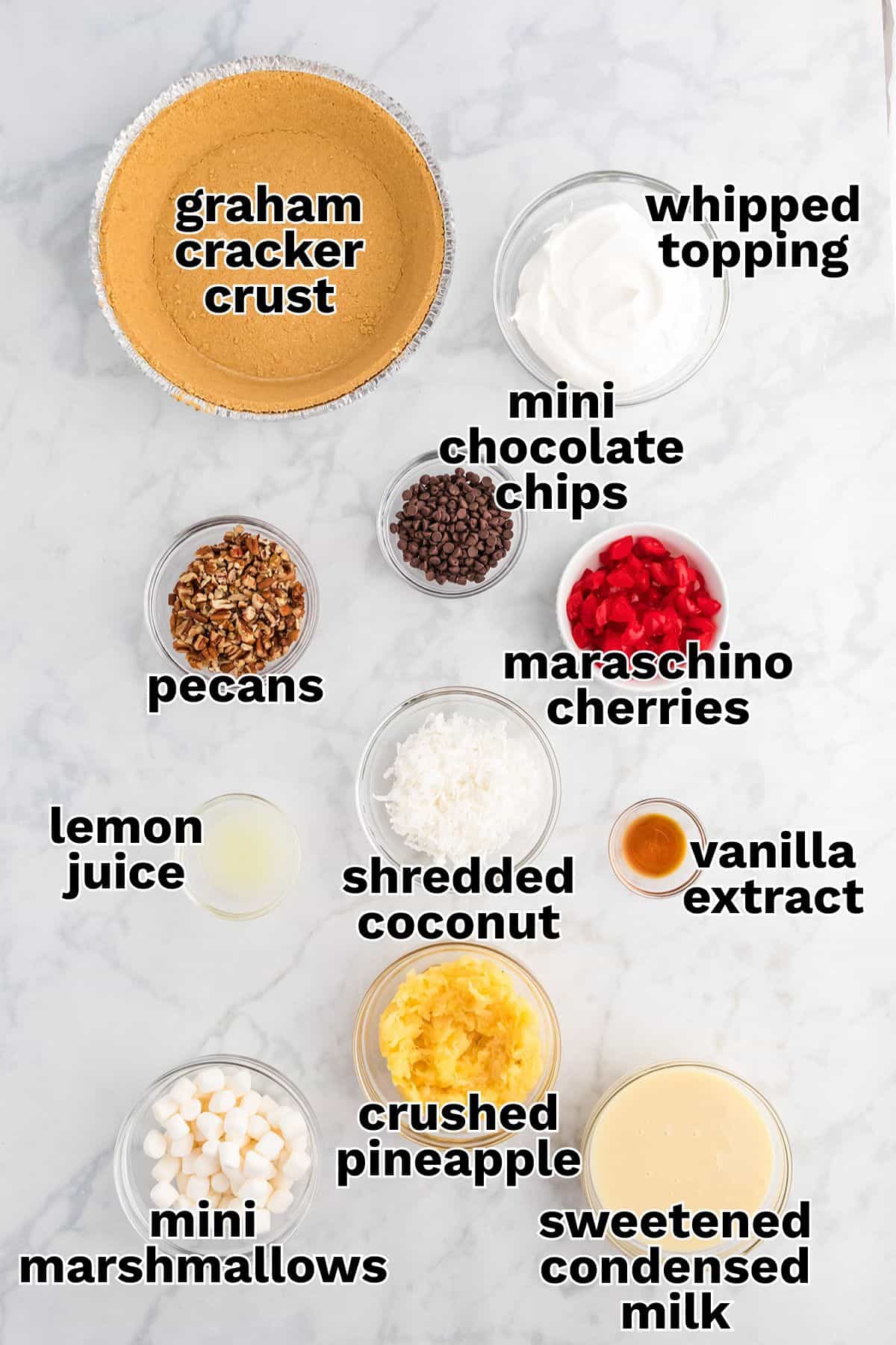 Ingredients for Million Dollar Pie dessert arranged on a marble surface, including graham cracker crust, whipped topping, mini chocolate chips, maraschino cherries, pecans, lemon juice, shredded coconut, crushed pineapple, mini marshmallows, vanilla extract, and sweetened condensed milk.