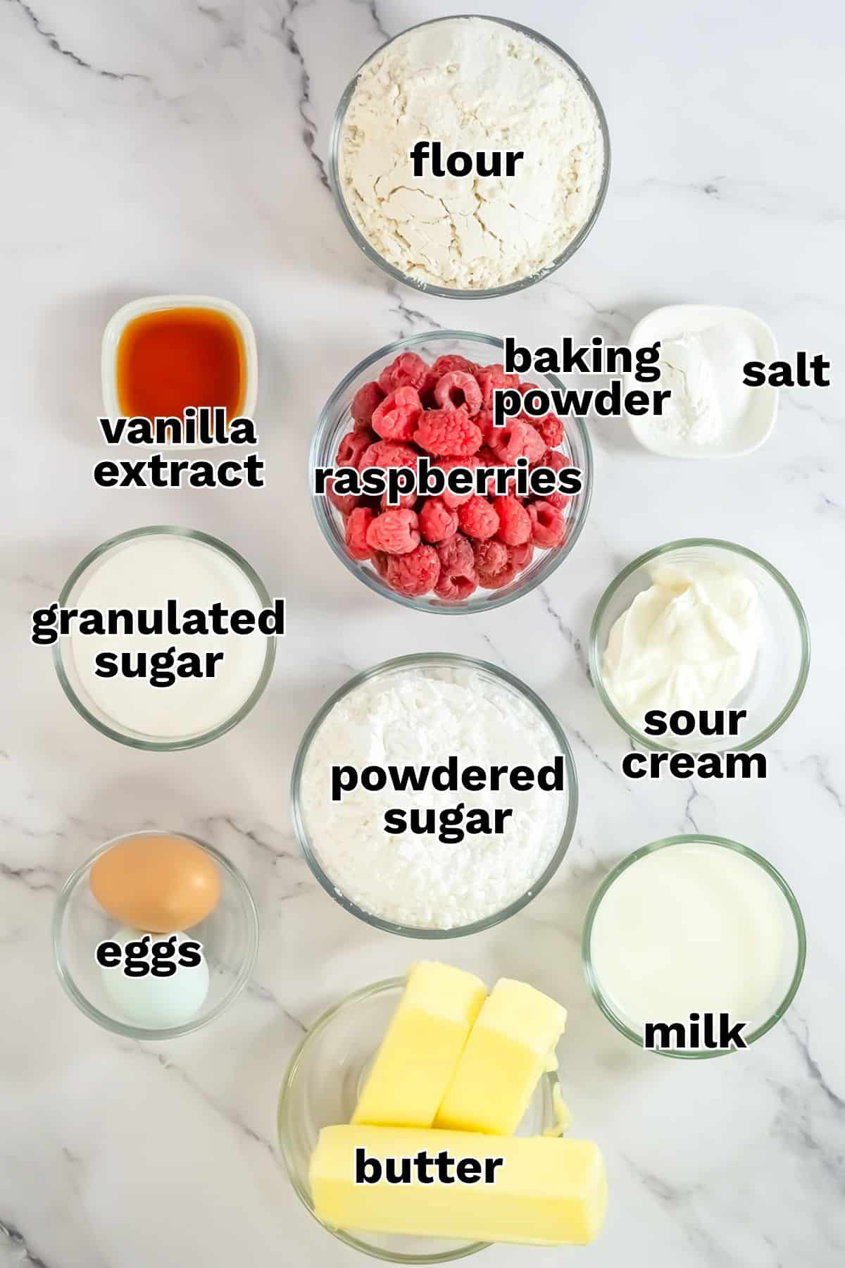 Top-down view of labeled baking ingredients in bowls for Raspberry Cake: flour, baking powder, salt, sour cream, milk, butter, eggs, granulated sugar, powdered sugar, vanilla extract, and raspberries.