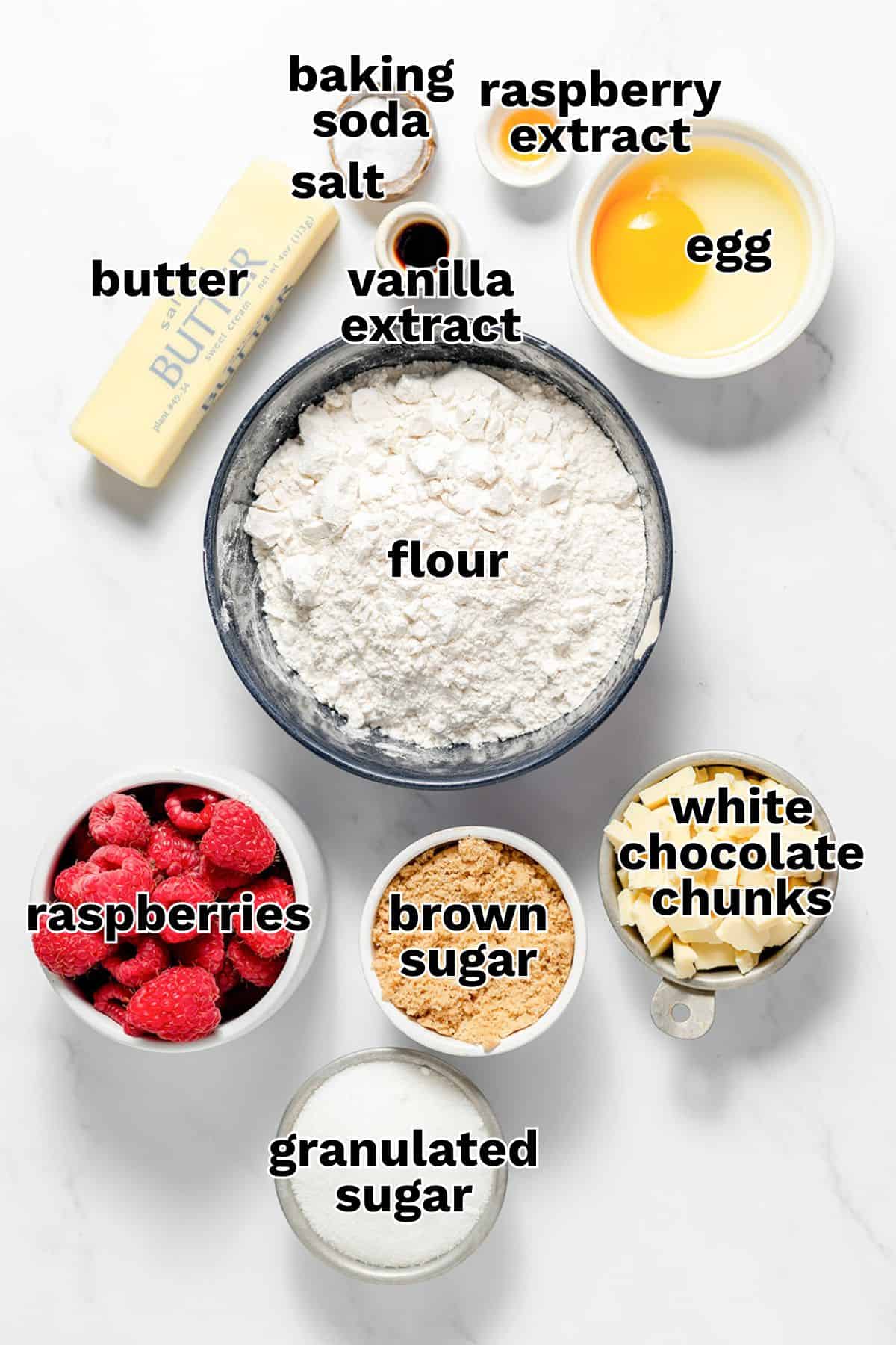 Baking ingredients for delicious Raspberry Cookies, including flour, butter, egg, vanilla and raspberry extract, baking soda, salt, raspberries, brown sugar, granulated sugar, and generous white chocolate chunks are arranged on a white surface.