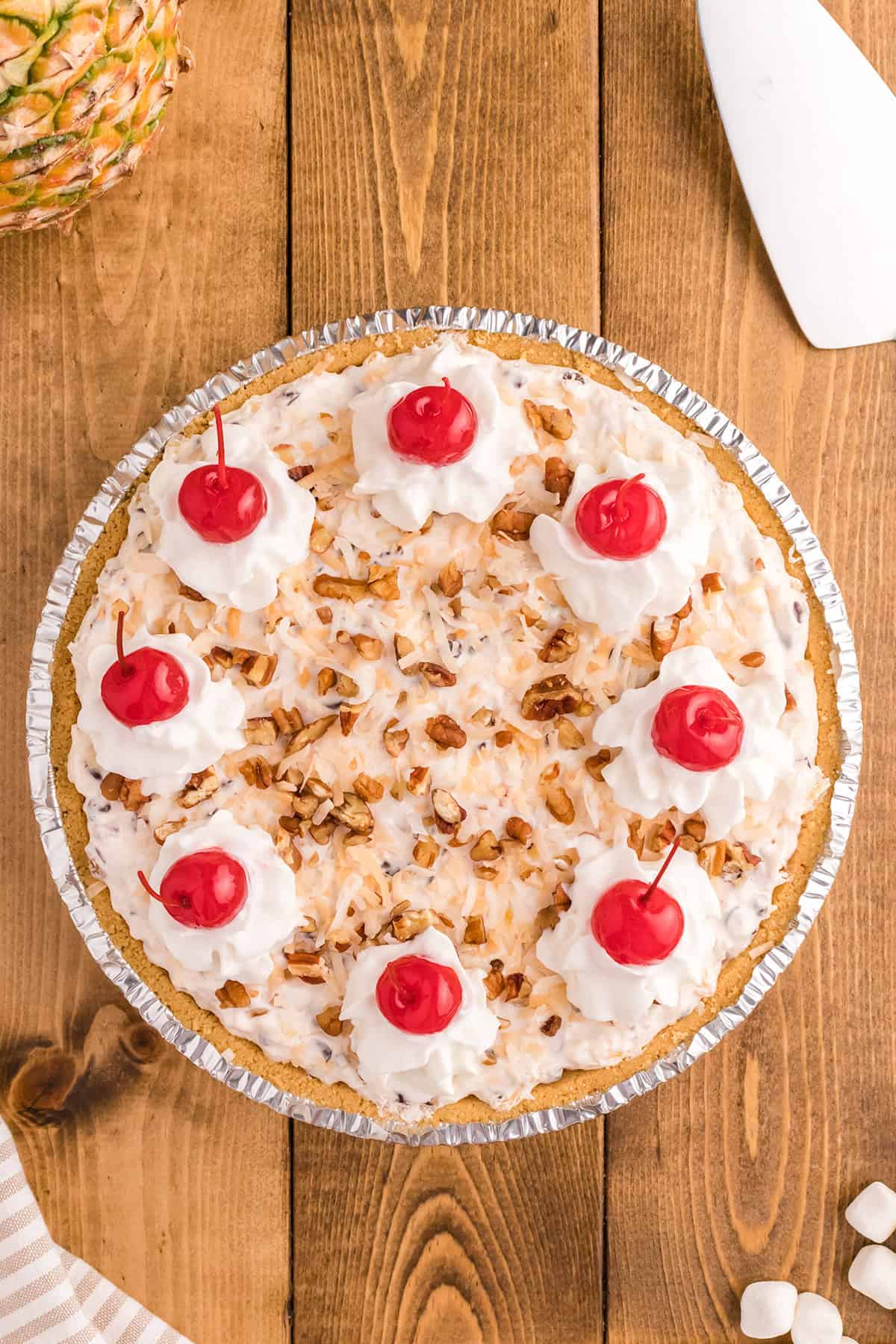 A Million Dollar Pie topped with whipped cream, chopped nuts, shredded coconut, and maraschino cherries is shown on a wooden surface next to a pie server.