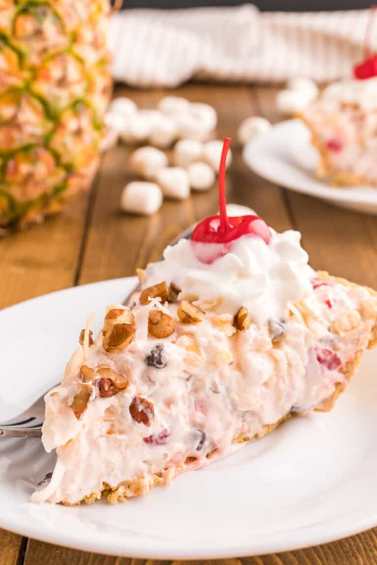 A slice of creamy Million Dollar Pie, topped with whipped cream and a cherry, served on a white plate with a fork. Pineapple and marshmallows are visible in the background.