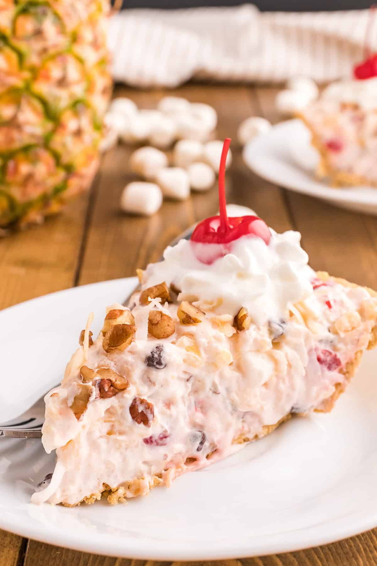 A slice of creamy Million Dollar Pie, topped with whipped cream and a cherry, served on a white plate with a fork. Pineapple and marshmallows are visible in the background.