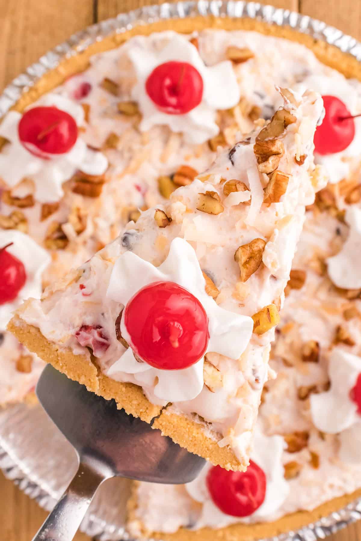 A slice of Million Dollar Pie with a crumb crust, creamy filling, whipped cream, chopped nuts, and a maraschino cherry on top is being lifted from the whole pie.