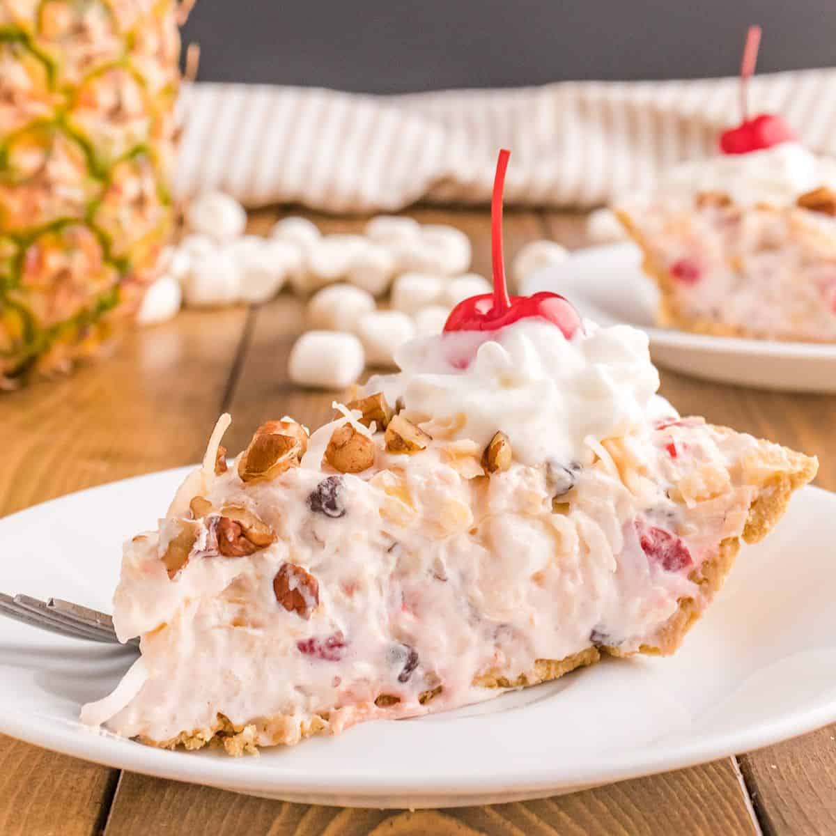 A slice of creamy Million Dollar Pie, this indulgent dessert is topped with whipped cream, a cherry, chopped nuts, and toasted coconut, served on a white plate.