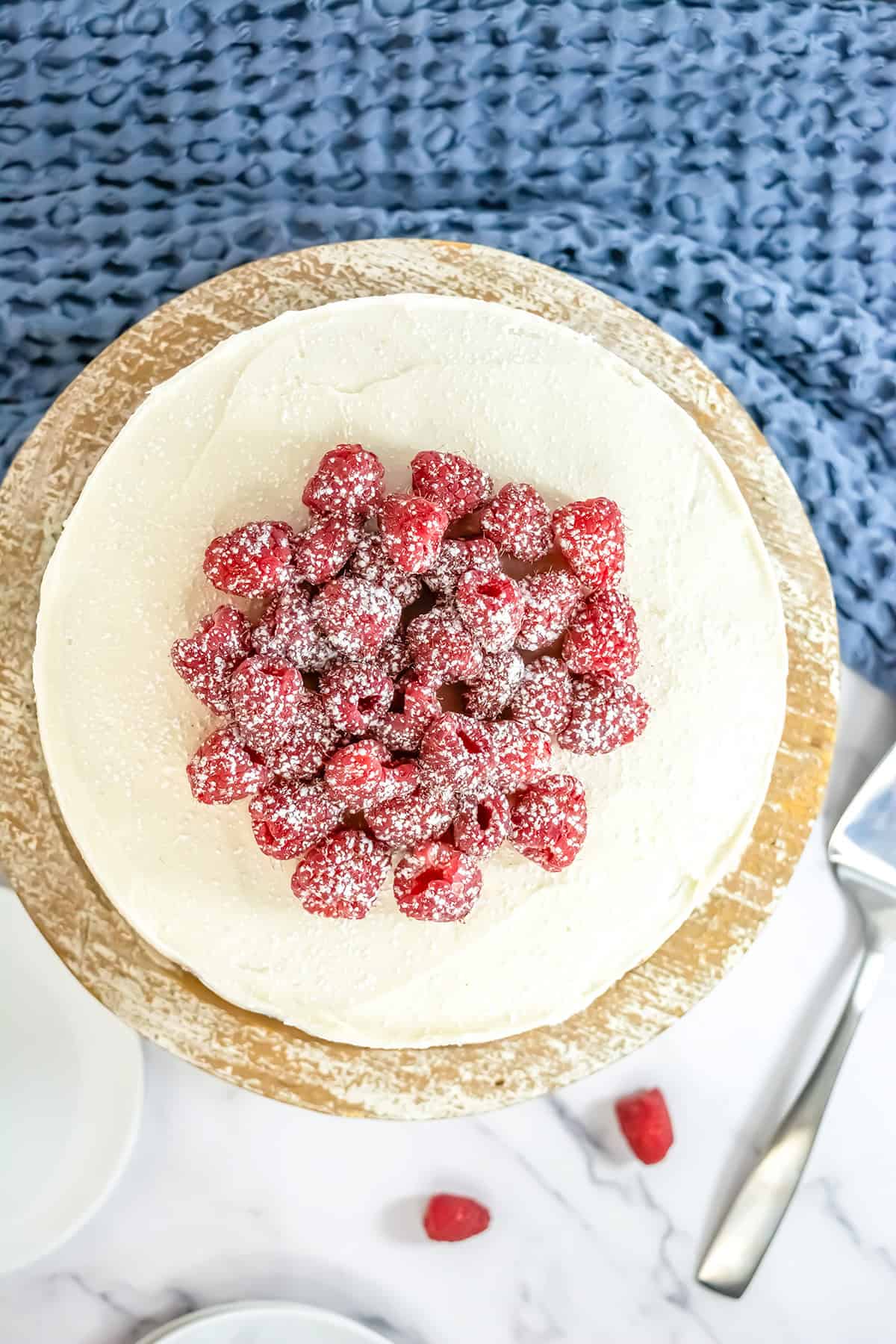 A round Raspberry Cake with white frosting, topped with fresh raspberries and powdered sugar, sits on a wooden stand. A cake server and a few raspberries are nearby on a marble surface.