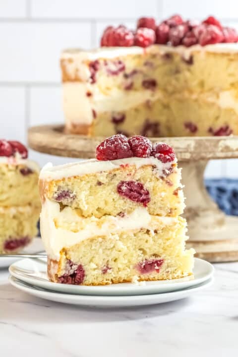 A delectable slice of layered raspberry cake with white frosting and raspberries on top is placed on a plate in front of the remaining Raspberry Cake resting elegantly on a stand.
