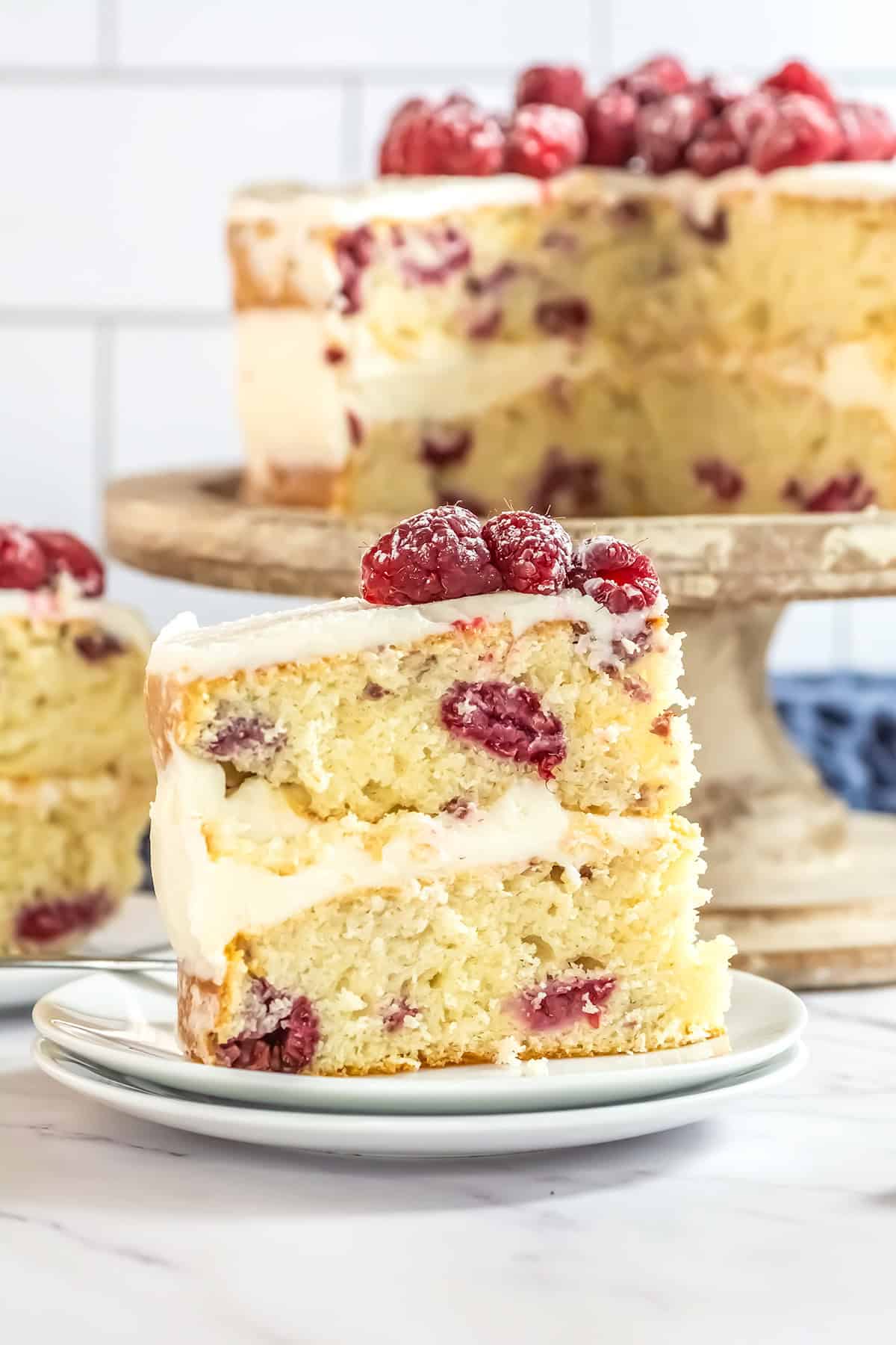 A delectable slice of layered raspberry cake with white frosting and raspberries on top is placed on a plate in front of the remaining Raspberry Cake resting elegantly on a stand.