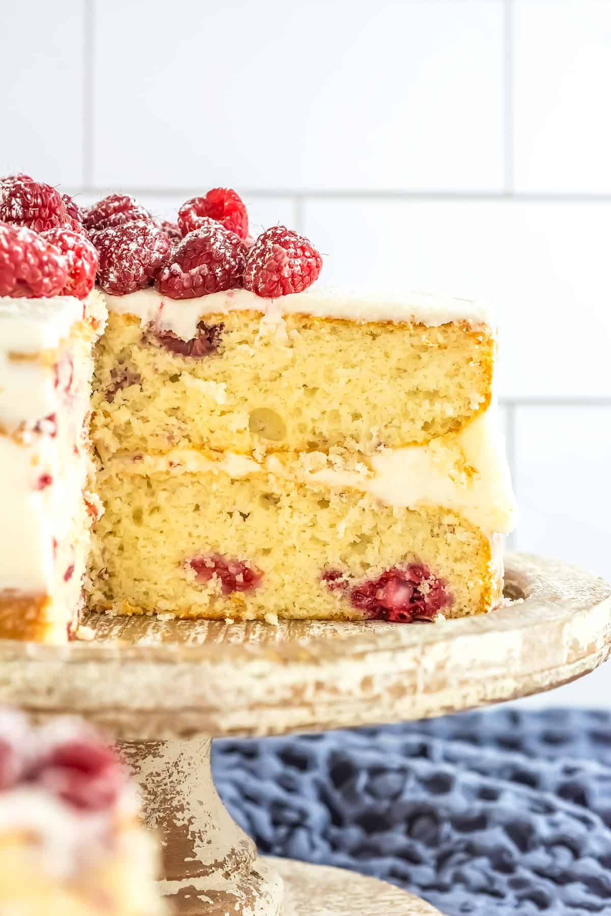 A Raspberry Cake featuring two layers with white frosting and fresh raspberries on top, shown sliced to reveal a fluffy interior studded with juicy berries, displayed on a rustic cake stand.