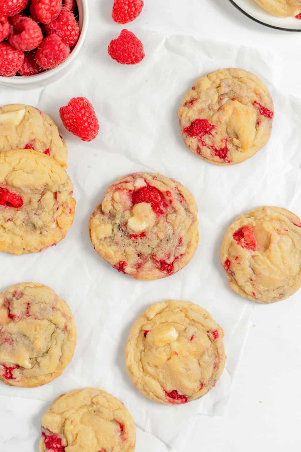 Seven Raspberry Cookies with visible raspberry pieces are arranged on white parchment paper, with loose raspberries and a bowl of raspberries nearby.
