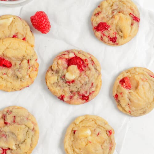 Seven Raspberry Cookies with visible raspberry pieces are arranged on white parchment paper, with loose raspberries and a bowl of raspberries nearby.