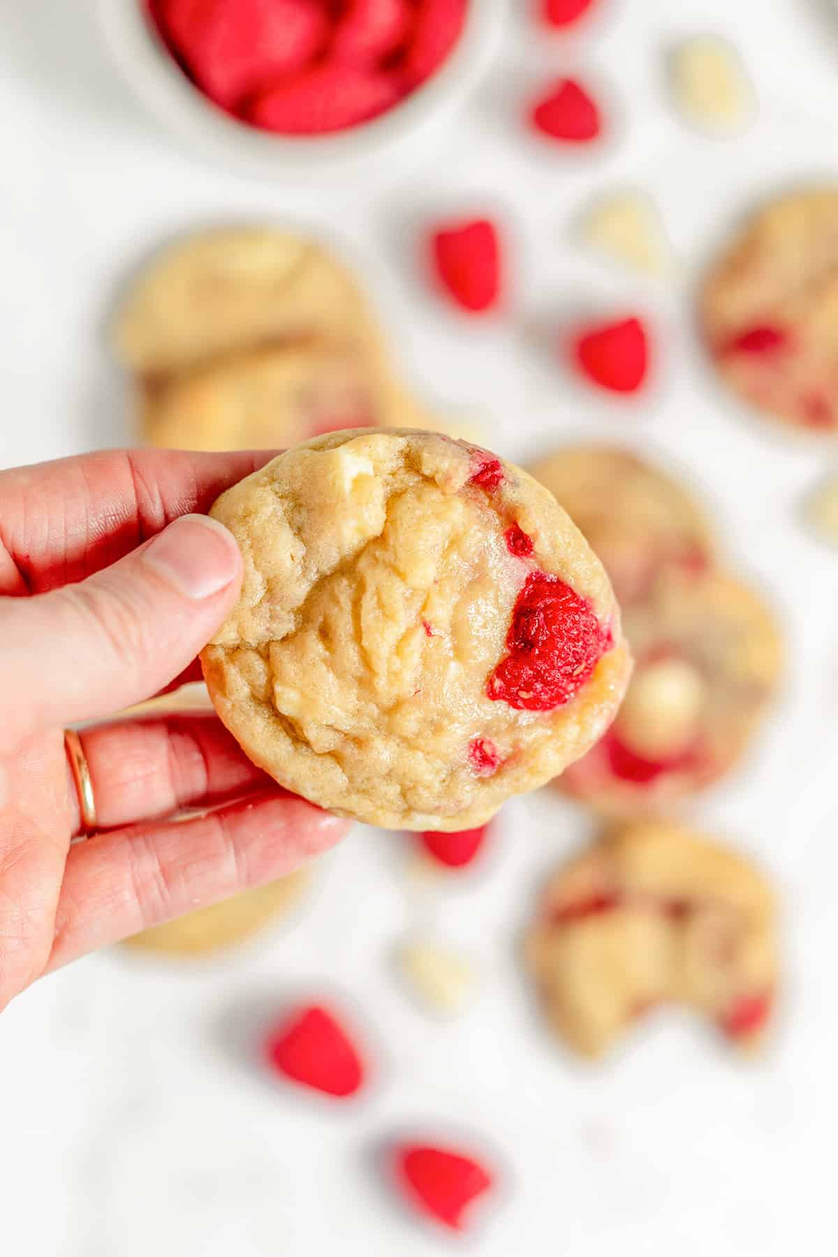 A hand holds a raspberry cookie studded with visible white chocolate chunks, while more white chocolate cookies and fresh raspberries are blurred in the background.