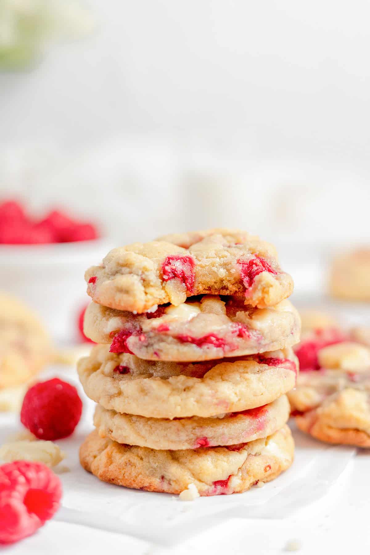 A stack of five White Chocolate Raspberry Cookies sits on white parchment paper, with fresh raspberries scattered around.